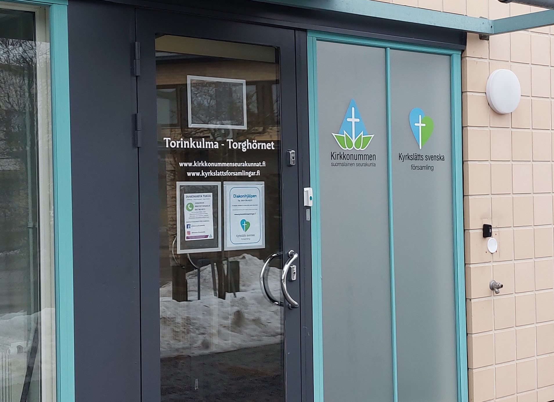 Lasinen sisäänkäyntiovi, jossa lukee tilan nimi, Torinkulma - Torghörnet. Oven viereisessä ikkunassa on Kirkkonummen seurakuntien sini-valko-vihreät logot.