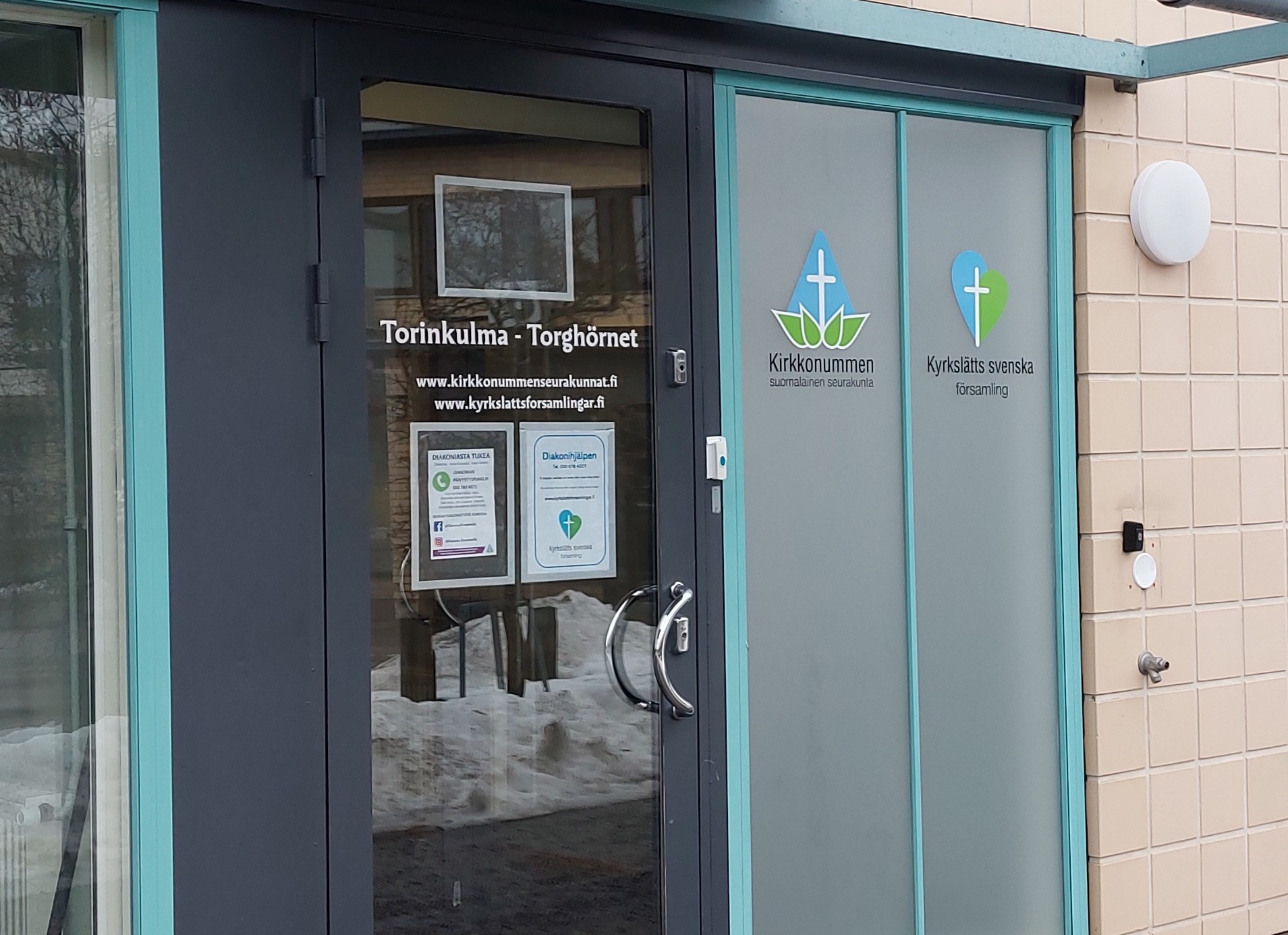 Torinkulma Lasinen sisäänkäyntiovi, jossa lukee tilan nimi, Torinkulma - Torghörnet. Oven viereisessä ikkunassa on Kirkkonummen seurakuntien sini-valko-vihreät logot.